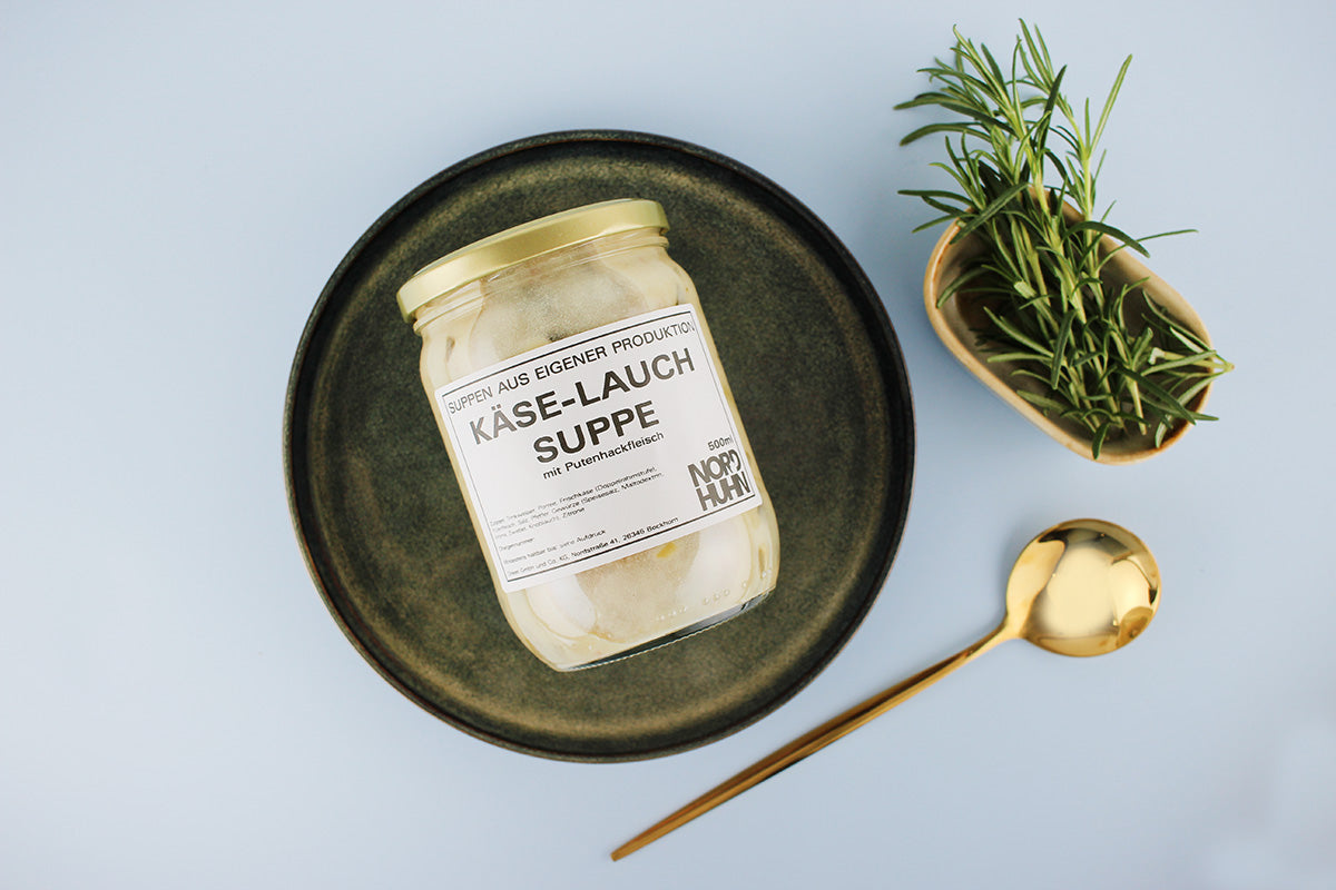 Ein Glas Käse-Lauch-Suppe mit Putenhackfleisch auf einem dunklen Teller, daneben ein goldener Löffel und ein kleines Gefäß mit frischem Rosmarin