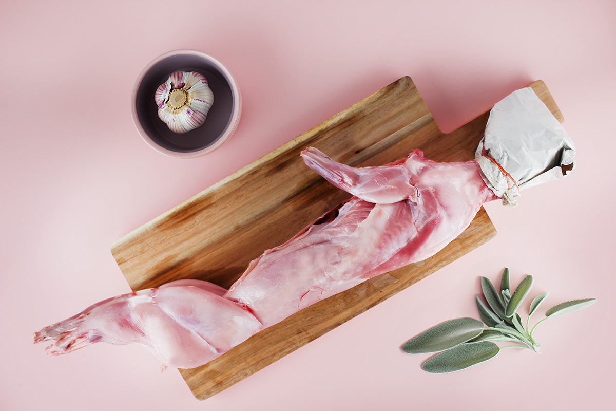 Ausgenommenes rohes Kaninchen auf einem Holzbrett, Knoblauch in einer Schale und frische Salbeiblätter auf rosa Untergrund