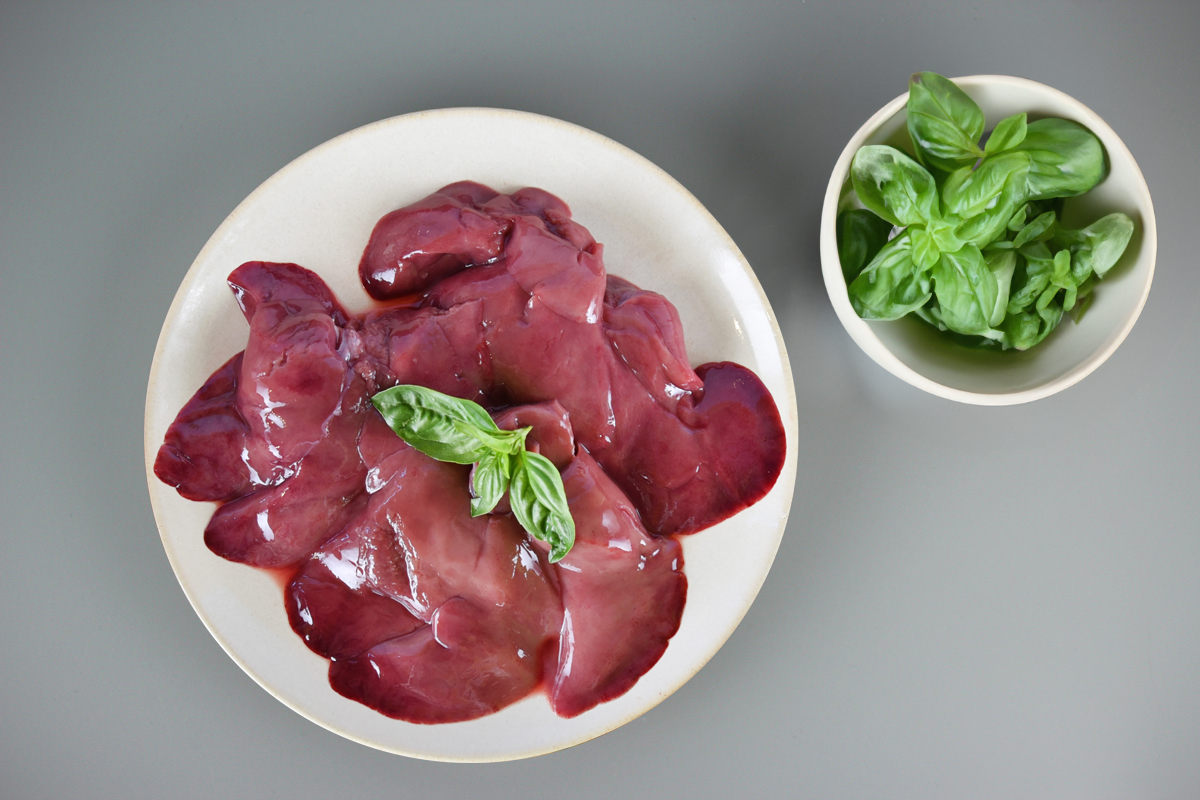 Rohe Kaninchenleber auf weißem Teller mit Basilikumblättern, daneben eine kleine Schale mit frischem Basilikum auf grauem Untergrund