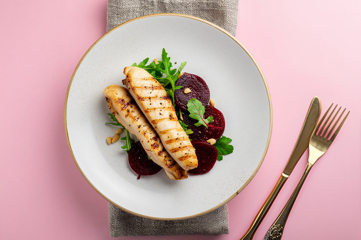 Zwei gegrillte Kaninchenfilets auf Rucola und Rote-Bete-Scheiben auf weißem Teller mit goldfarbenem Besteck auf rosa Untergrund