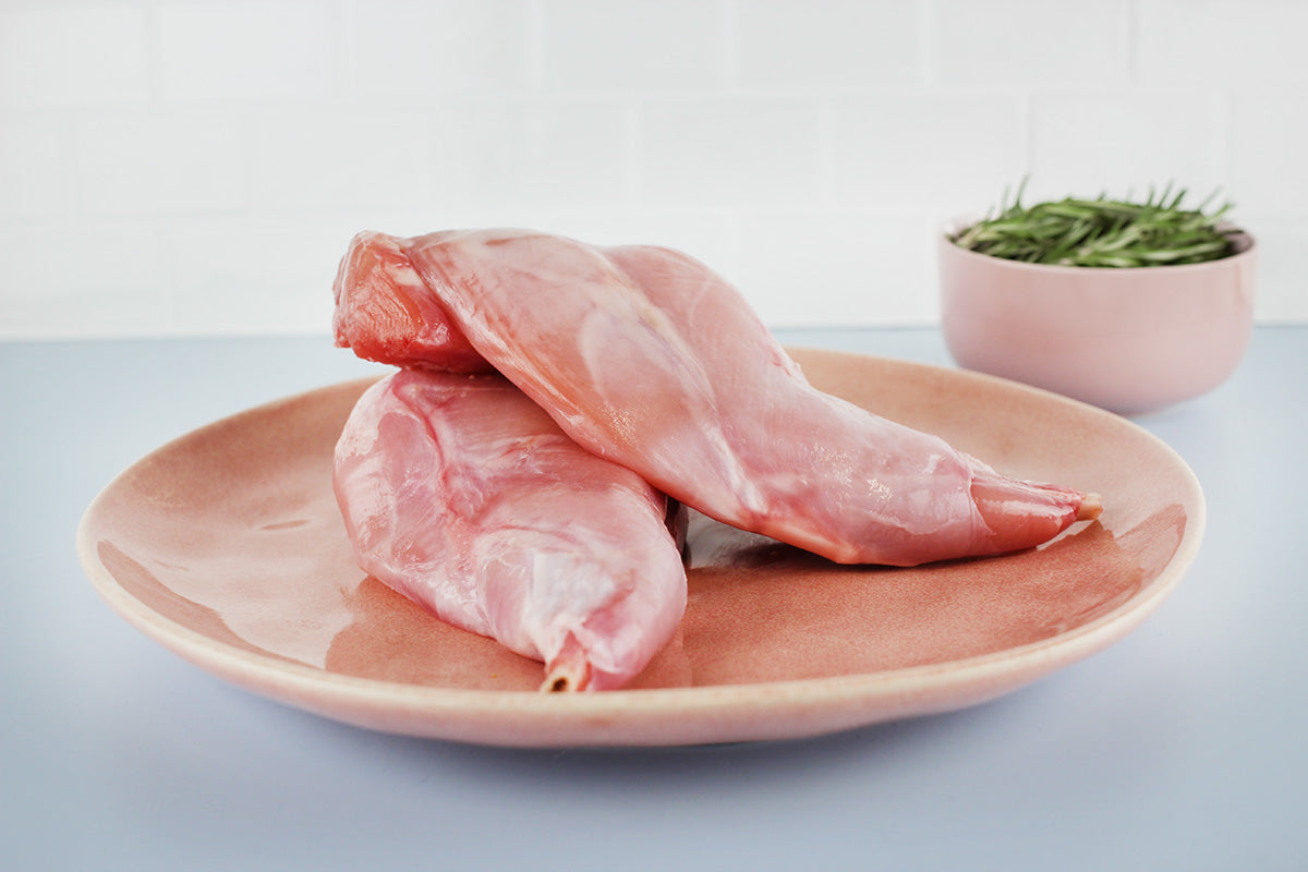 Zwei rohe Kaninchenkeulen auf einem runden, braunen Teller mit einer kleinen Schale frischer Kräuter im Hintergrund