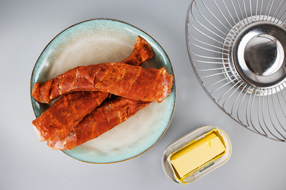 Zwei mit Speck umwickelte, gewürzte Kaninchenfilets auf einem blauen Teller, daneben Butter in einer Schale und ein Metallgitterkorb