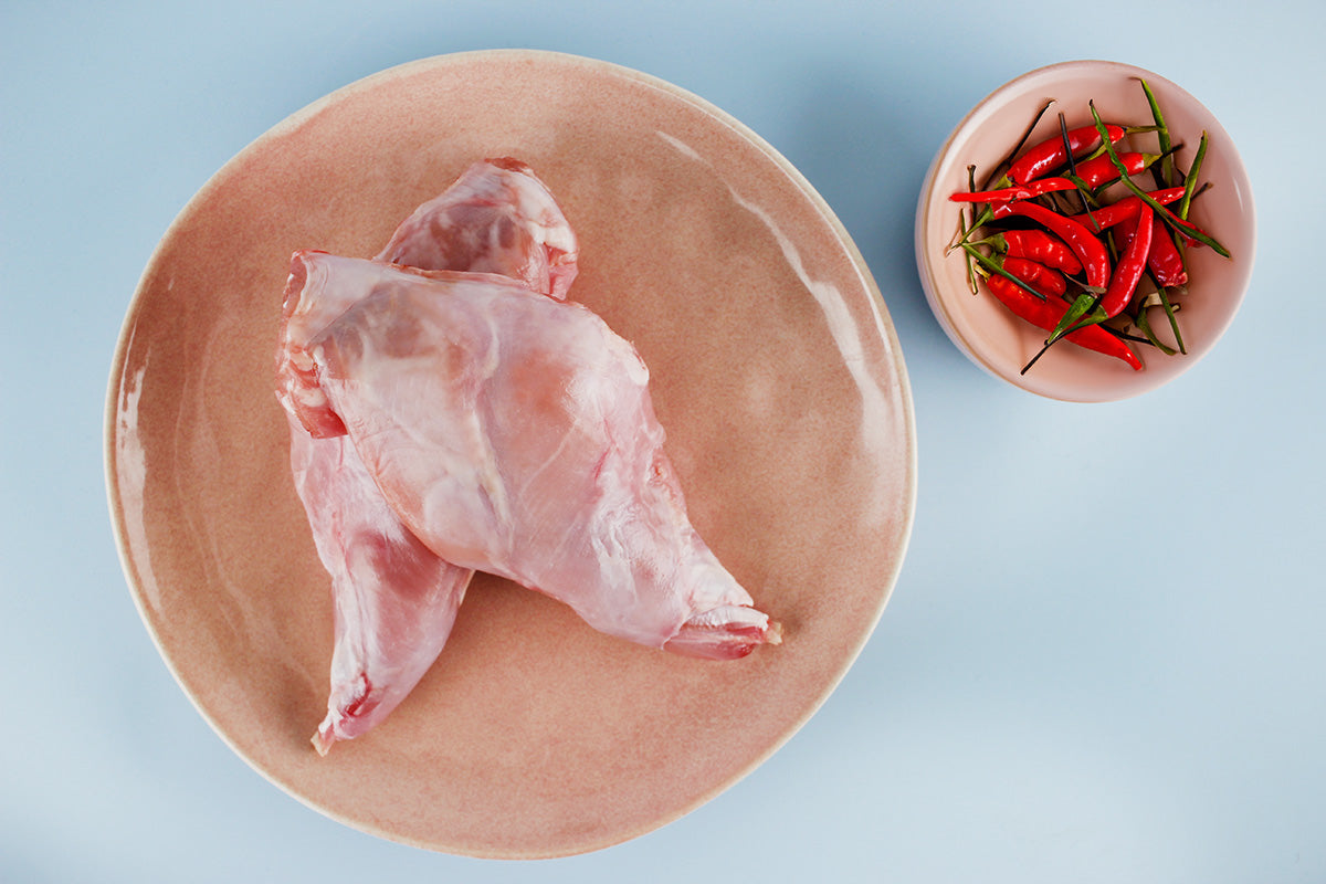 Zwei rohe Kaninchenkeulen auf einem braunen Teller neben einer kleinen Schale mit roten Chilischoten auf hellblauem Hintergrund