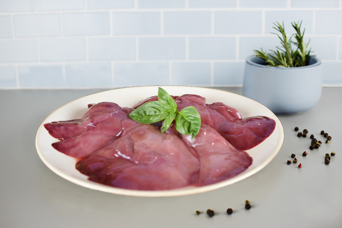 Rohe Kaninchenleber auf weißem Teller, garniert mit frischem Basilikum, daneben Pfefferkörner und ein Topf mit Rosmarin