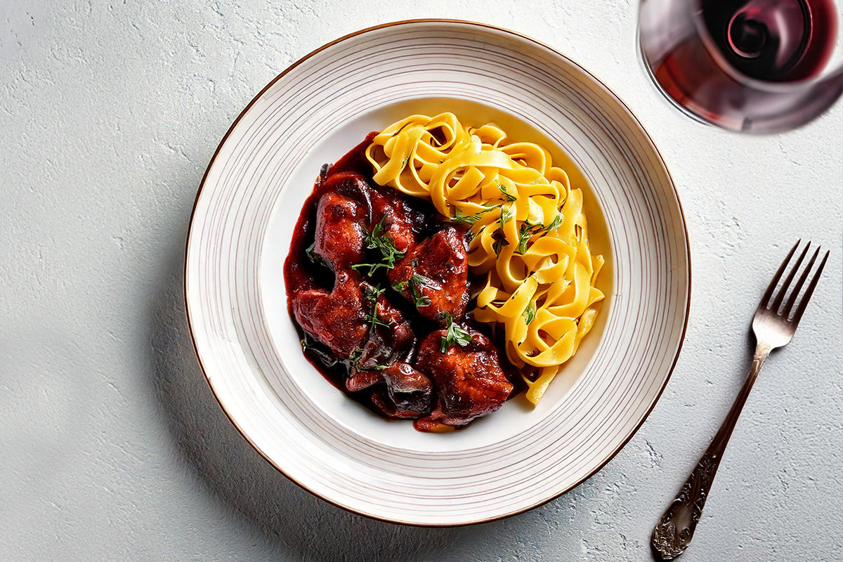 Kaninchenleber in Rotweinsauce serviert mit frischer Tagliatelle und Thymian auf weißem Teller, daneben Gabel und Glas Rotwein