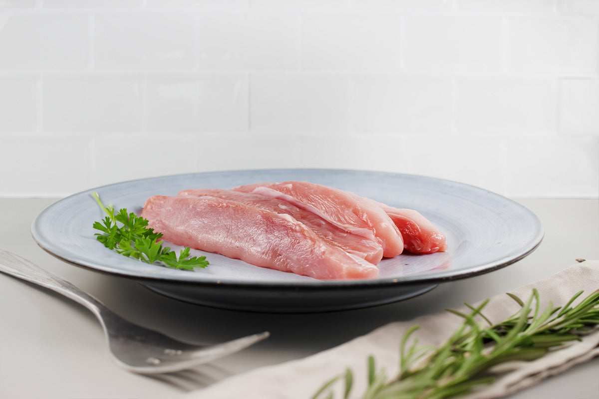 Drei rohe Kaninchenrückenfilets auf einem grauen Teller mit Petersilienzweig, daneben Gabel und Rosmarinzweig auf hellem Untergrund