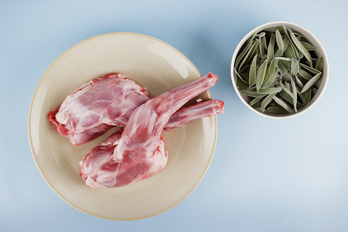 Zwei rohe Kaninchenvorderläufe auf einem hellen Teller neben einer Schale mit frischen Salbeiblättern auf blauem Hintergrund