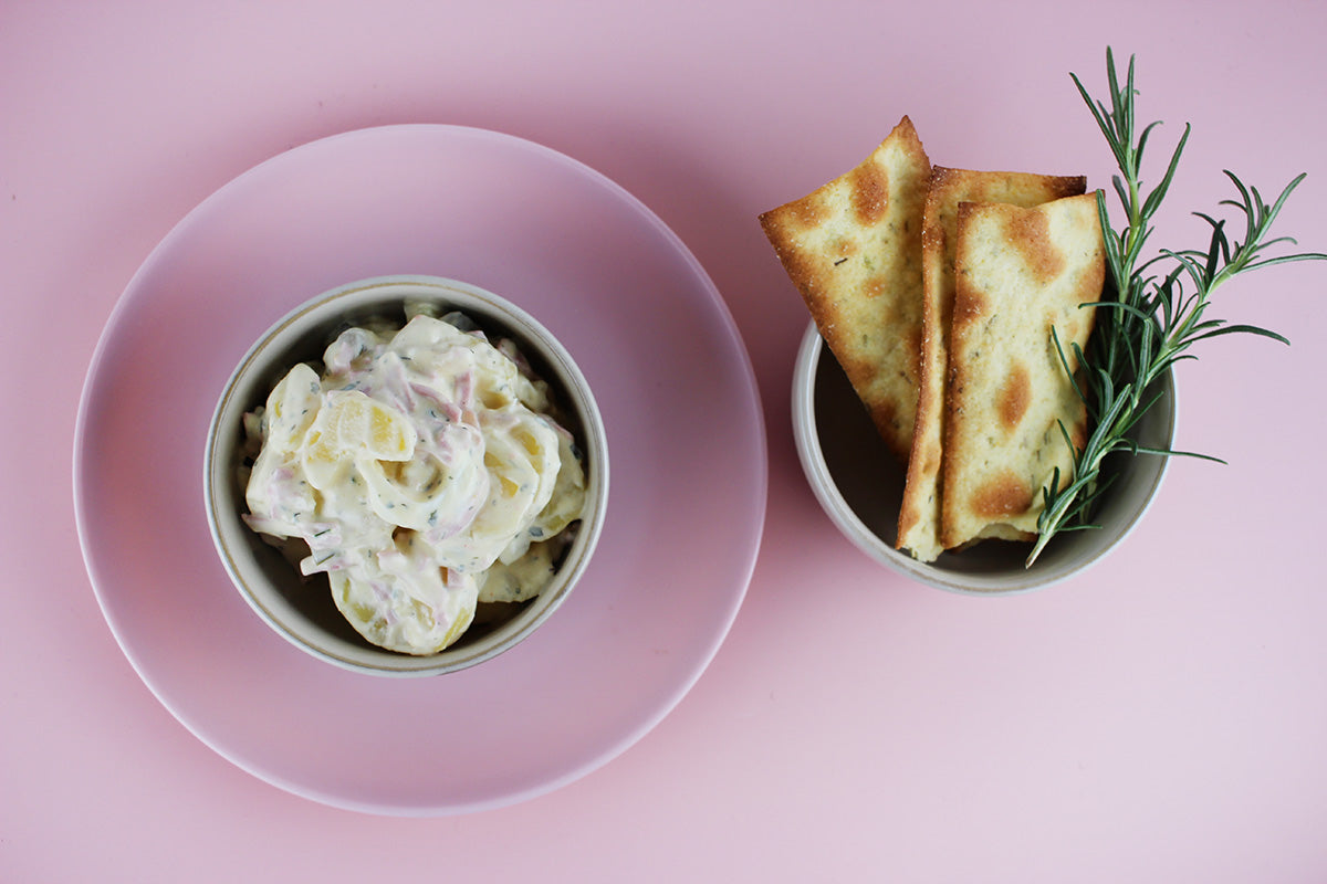 Kartoffelsalat mit Geflügelfleischwurst in einer Schale auf rosa Teller, daneben Fladenbrot mit Rosmarinzweig in kleiner Schale