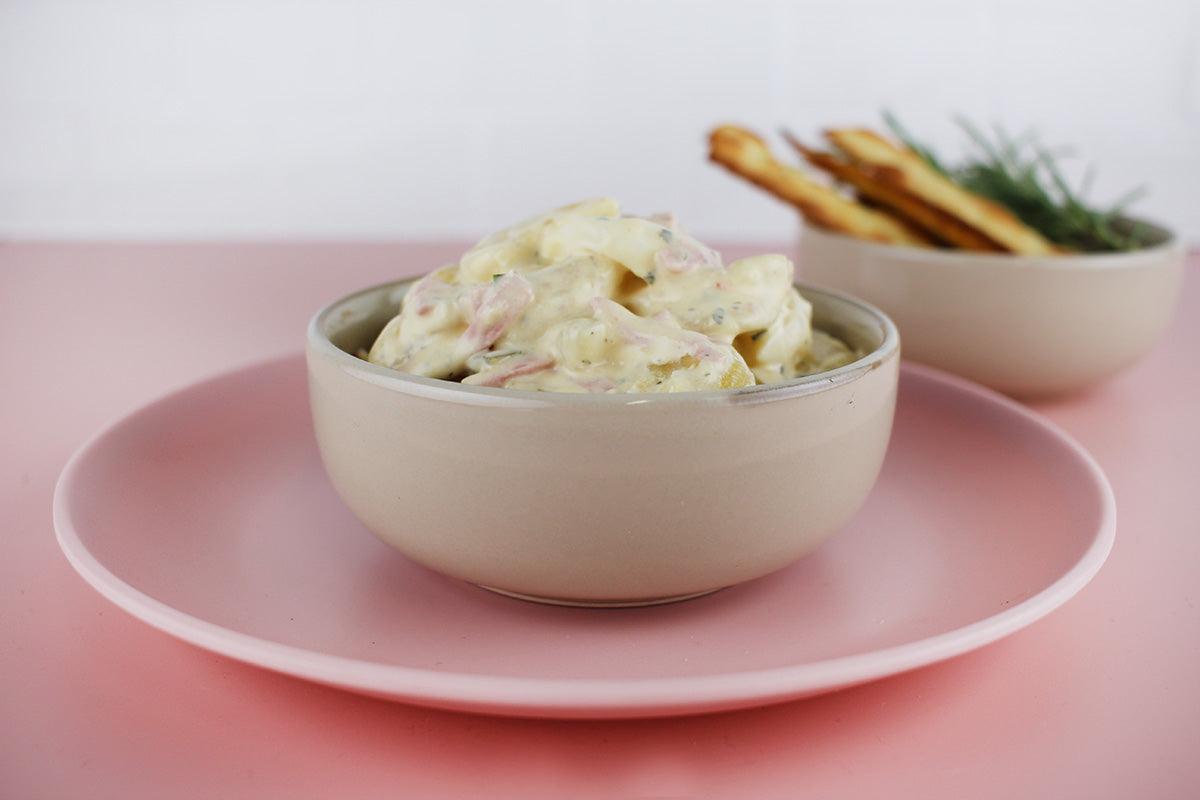 Schüssel mit cremigem Kartoffelsalat mit Geflügelfleischwurst auf rosa Teller, im Hintergrund Schale mit Brotsticks und Kräutern