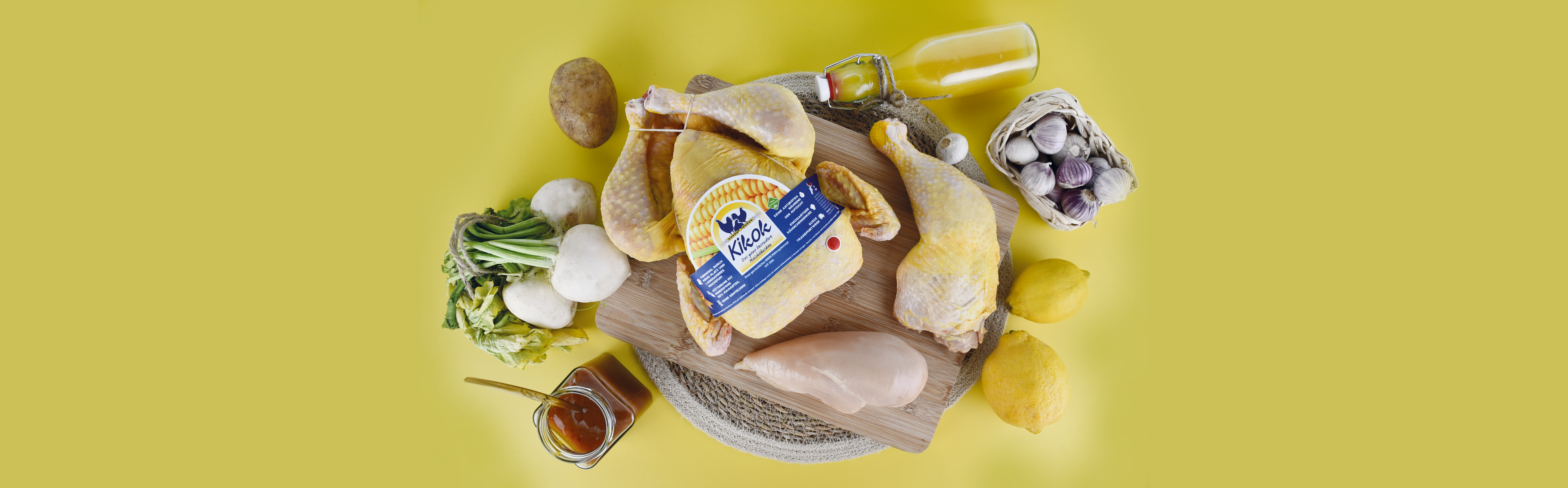 Rohe Kikok-Hähnchenteile auf Holzbrett mit Honig, Knoblauch, Kartoffel, Zitronen und grünem Gemüse auf gelbem Hintergrund