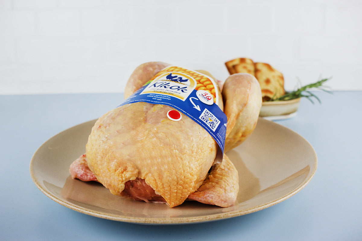 Ungekochtes Kikok Brathähnchen mit blauer Verpackung auf einem beigefarbenen Teller, im Hintergrund Brot und Rosmarin