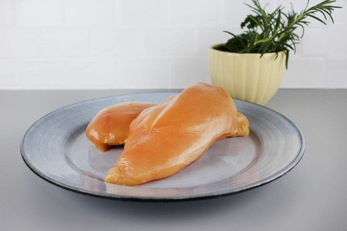 Zwei rohe Kikok Hähnchenbrustfilets auf einem grauen Teller, im Hintergrund ein gelber Topf mit frischen Kräutern