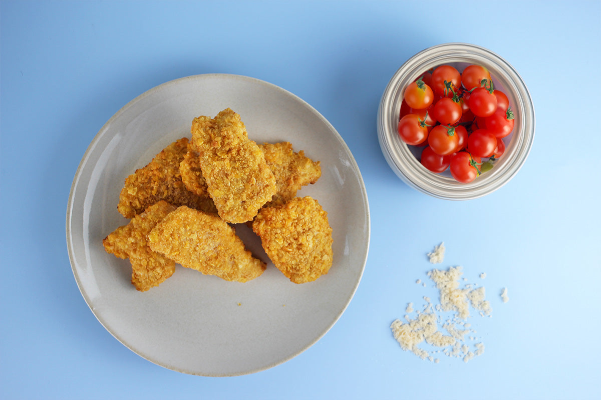 Sechs knusprige Kikok Chicken Nuggets auf einem grauen Teller, daneben eine Schale mit roten Kirschtomaten und etwas Paniermehl auf blauem Untergrund