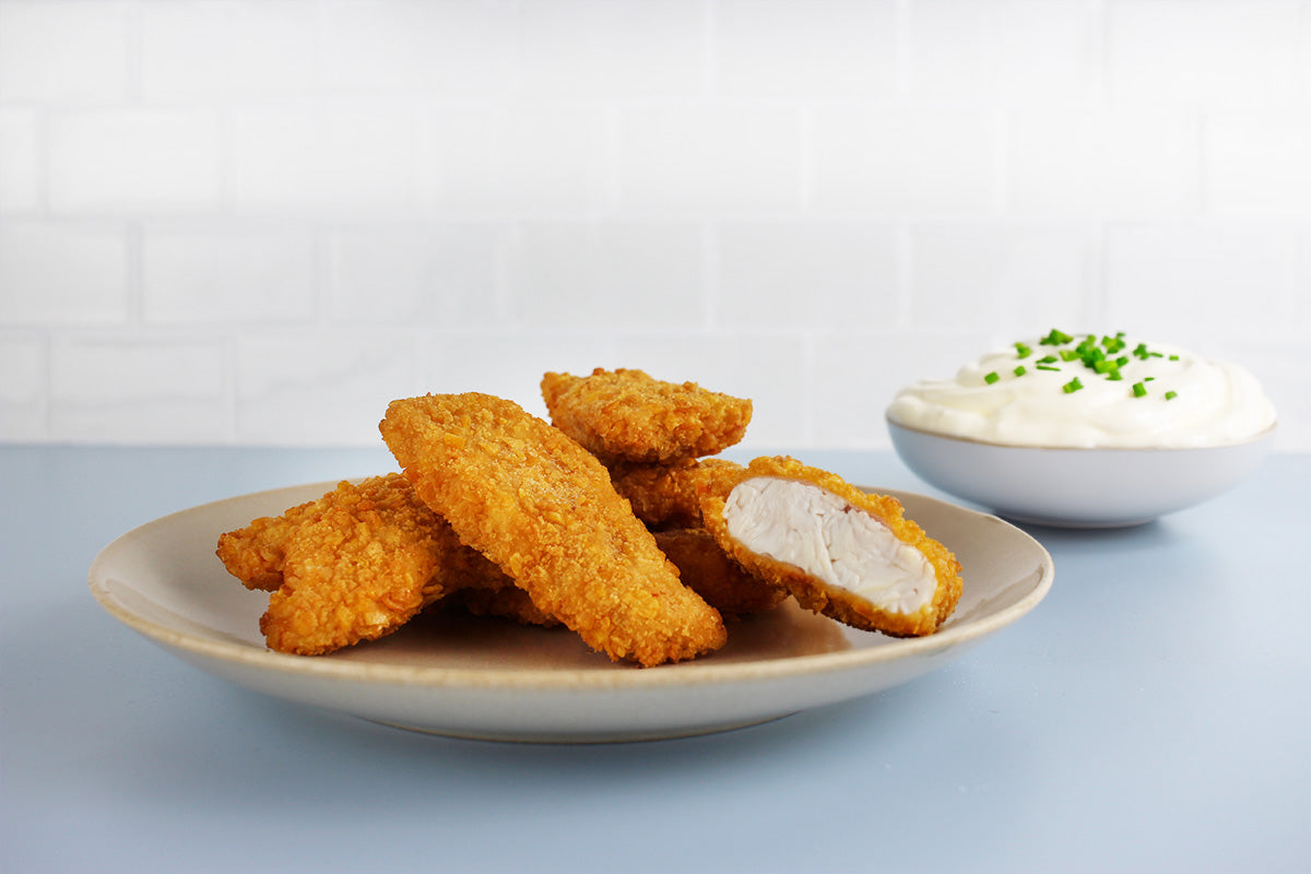 Mehrere goldbraune Kikok Chicken Nuggets auf einem weißen Teller, eine halbierte Nugget zeigt das weiße Hühnerfleisch, daneben eine Schale mit weißer Sauce und grünen Kräutern