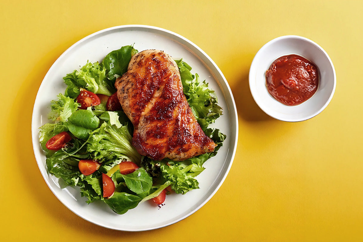 Gegrilltes Hähnchensteak auf gemischtem grünen Salat mit Tomaten, daneben Schale mit roter BBQ-Sauce auf gelbem Hintergrund