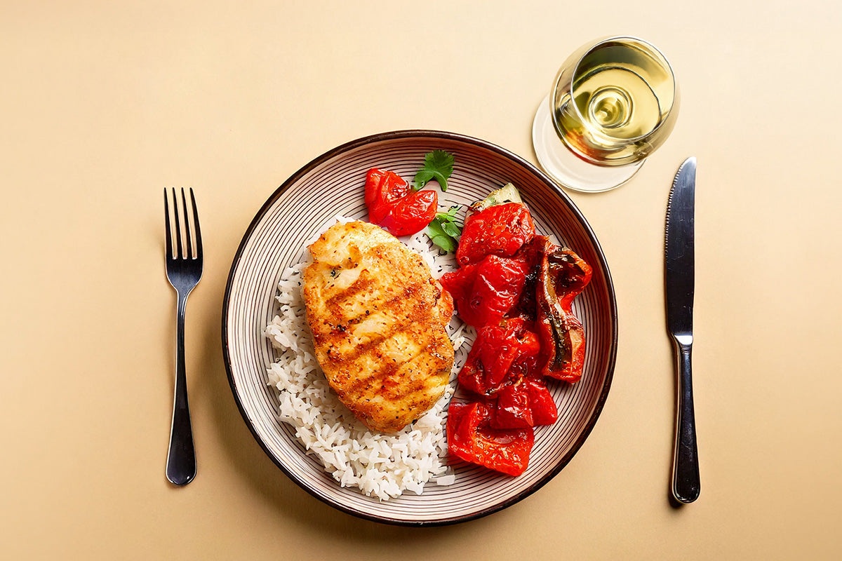 Gegrilltes Hähnchensteak auf weißem Reis mit gegrillten roten Paprikastücken auf einem Teller, daneben Besteck und ein Glas Weißwein