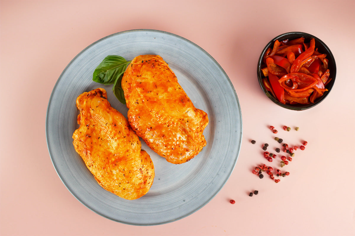 Zwei Hähnchensteaks mit Paprikamarinade auf grauem Teller, daneben Schale mit Paprikastreifen und bunte Pfefferkörner auf rosa Untergrund