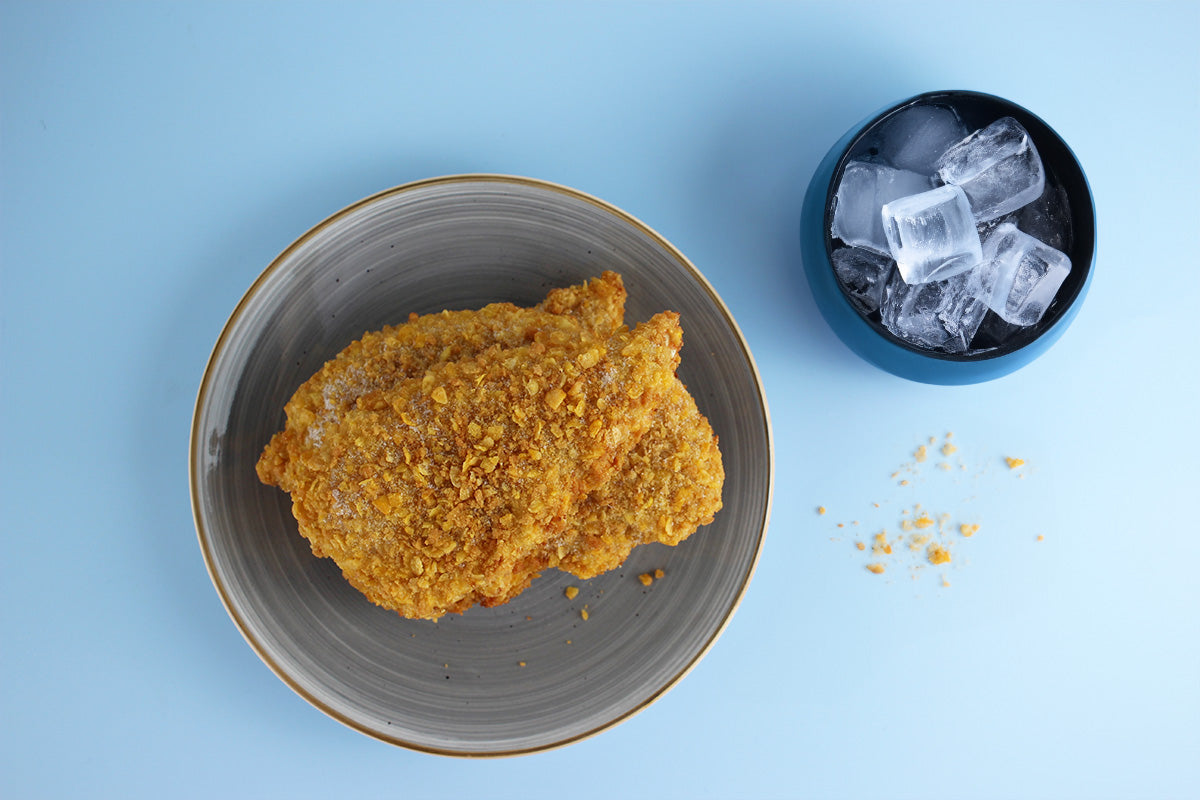 Zwei panierte Mini-Schnitzel auf grauem Teller neben schwarzer Schale mit Eiswürfeln auf blauem Hintergrund