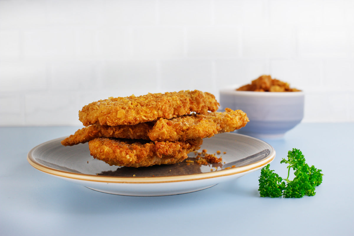 Drei knusprige, goldbraune Mini-Schnitzel auf einem grauen Teller mit einem kleinen Bund Petersilie und einer Schale mit Nuggets im Hintergrund