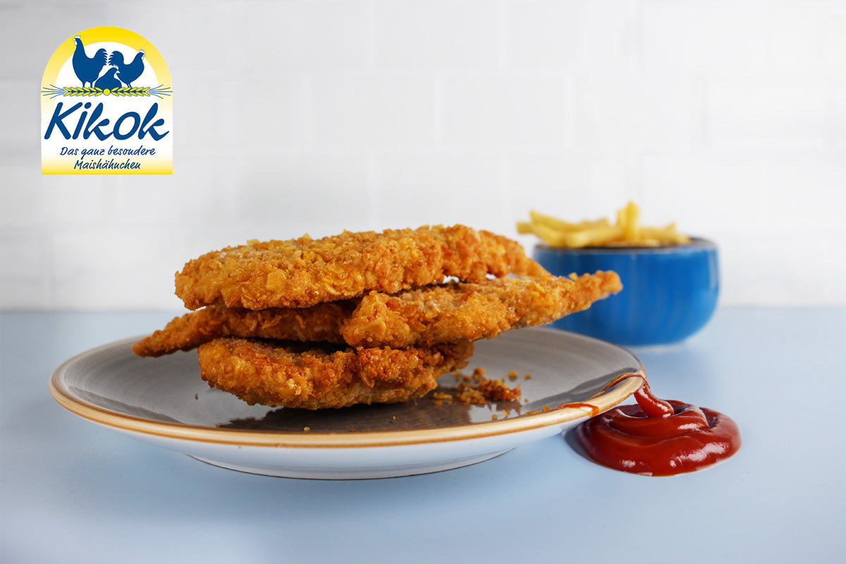 Drei goldbraun panierte Schnitzel auf einem Teller, daneben ein Klecks Ketchup und eine Schale mit Pommes. Oben links das Kikok-Logo mit zwei blauen Vögeln.