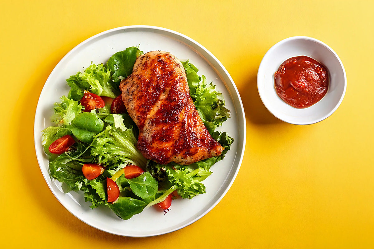 Gegrilltes Kikok-Hähnchensteak auf gemischtem grünen Salat mit Kirschtomaten, daneben kleine Schale mit roter Sauce auf gelbem Hintergrund