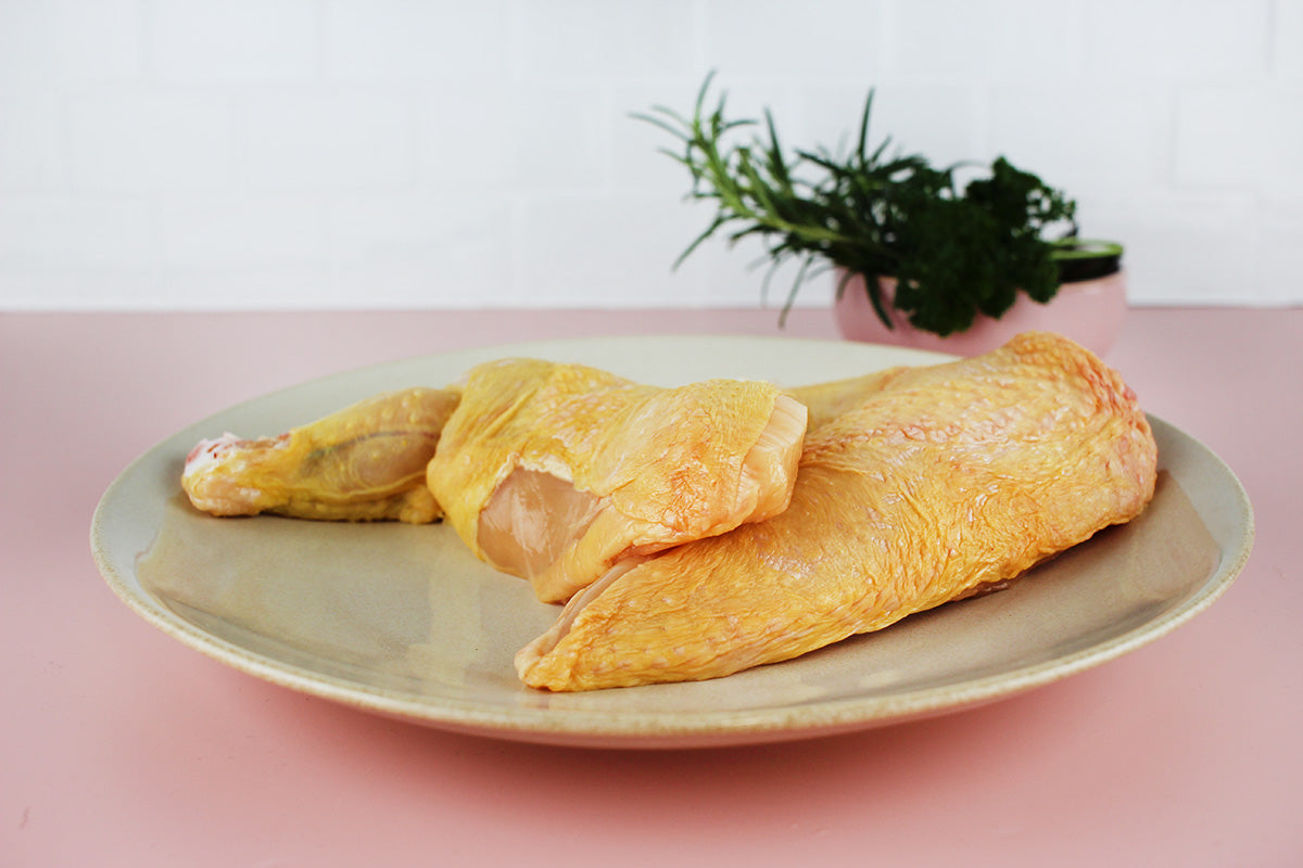 Rohe Hähnchenschenkel mit Haut auf einem hellen Teller, im Hintergrund frische Kräuter in einem Topf