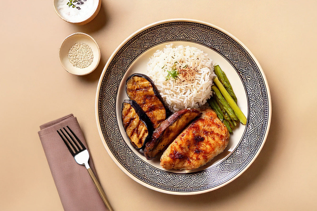 Gegrilltes mariniertes Hähnchenbrustfilet mit Reis, gegrillter Aubergine und grünem Spargel auf dekorativem Teller. Daneben eine Gabel auf Servierte und eine Schale mit Sesam und eine Schale mit Joghurt