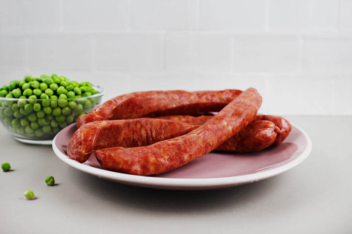 Drei lange, gebogene Kochwürste auf einem weißen Teller neben einer Glasschale mit grünen Erbsen