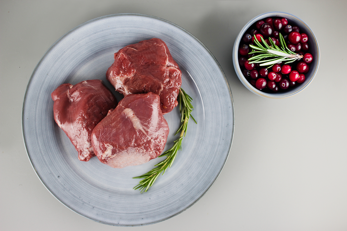 Drei rohe Lammhüften auf grauem Teller mit Rosmarinzweig, daneben Schale mit frischen Cranberries und Rosmarinzweig
