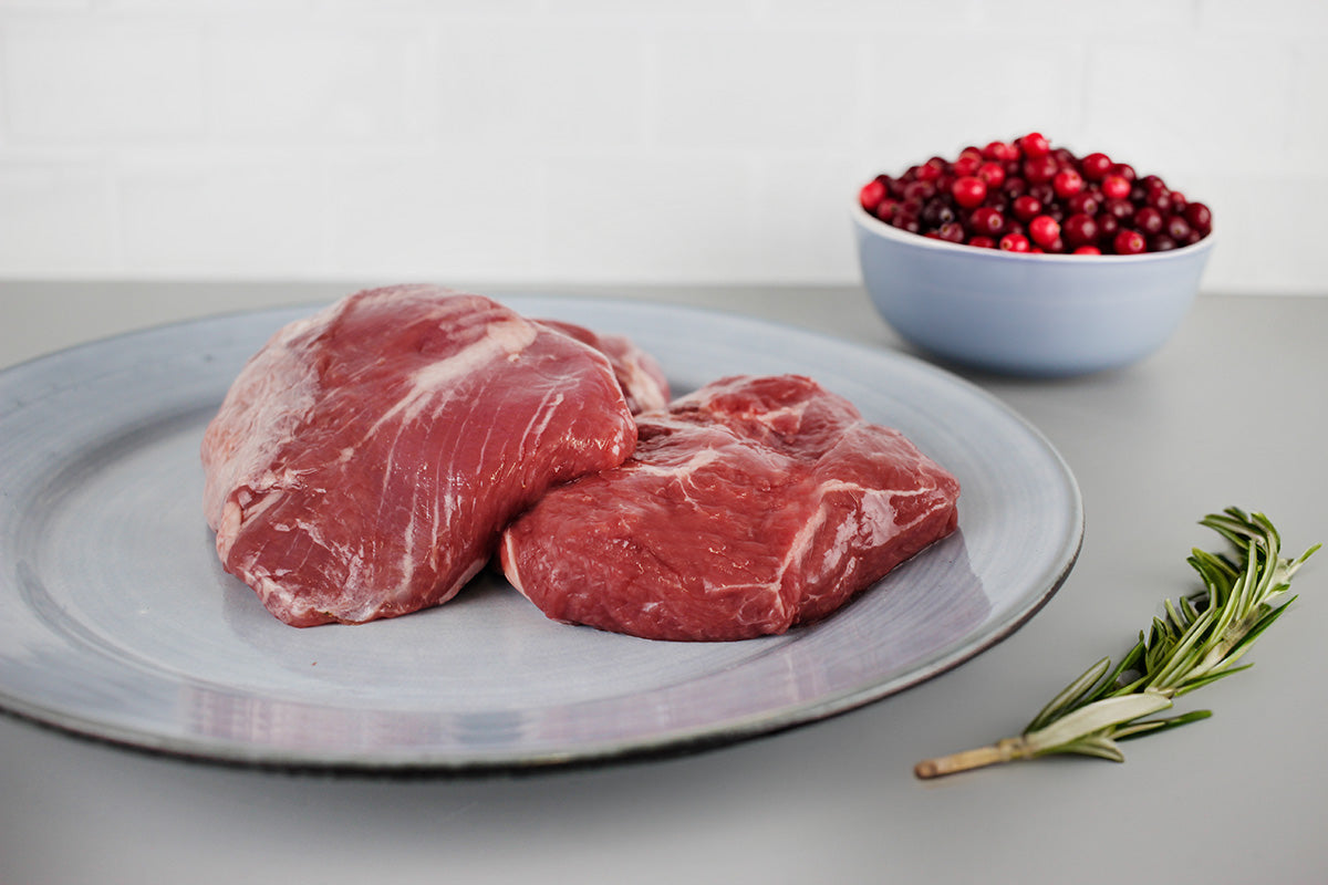 Zwei rohe Lammhüftsteaks auf einem grauen Teller, daneben eine Schale mit roten Beeren und ein Rosmarinzweig