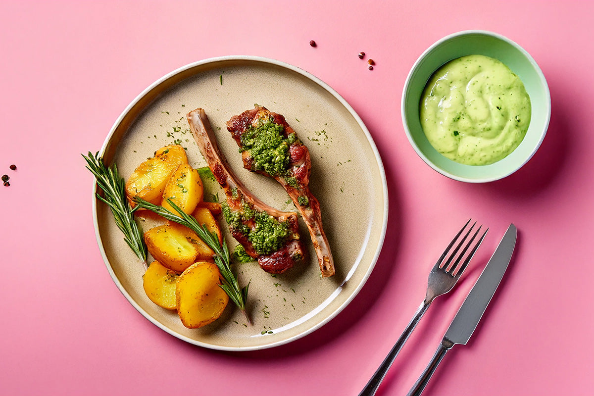 Zwei marinierte Lammkoteletts mit grüner Kräutersoße, gebratene Kartoffeln und Rosmarinzweige auf beigem Teller, daneben grüne Soße in Schale, Besteck auf rosa Untergrund