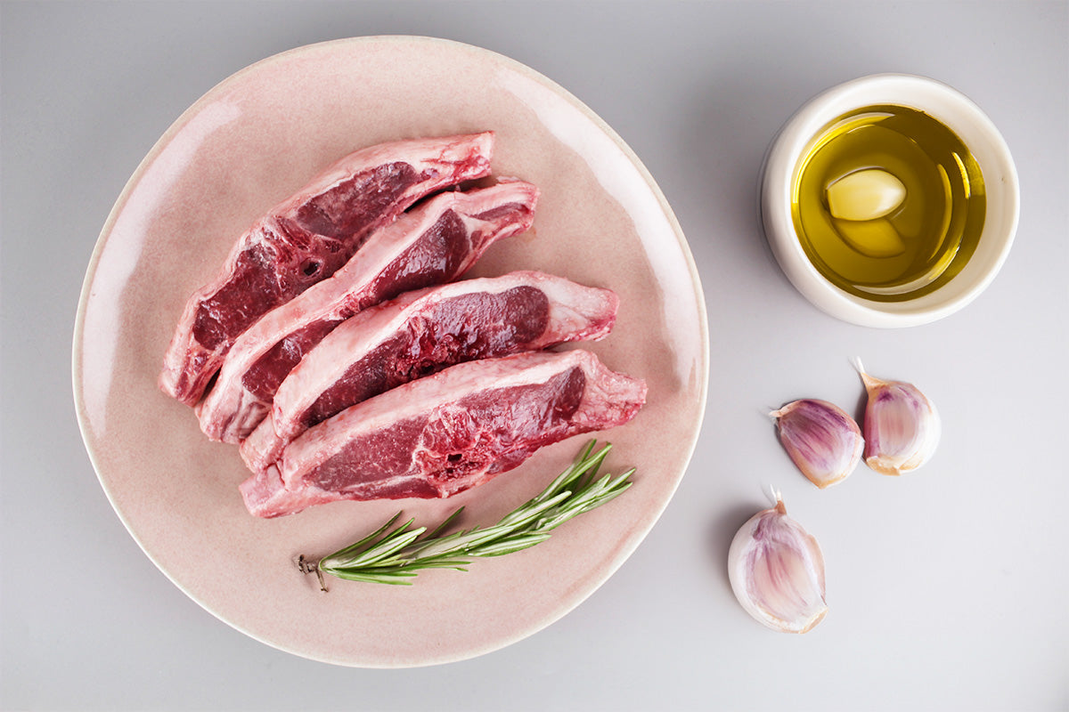 Vier rohe Lammkoteletts auf einem Teller mit Rosmarinzweig, daneben Knoblauchzehen und eine Schale Olivenöl