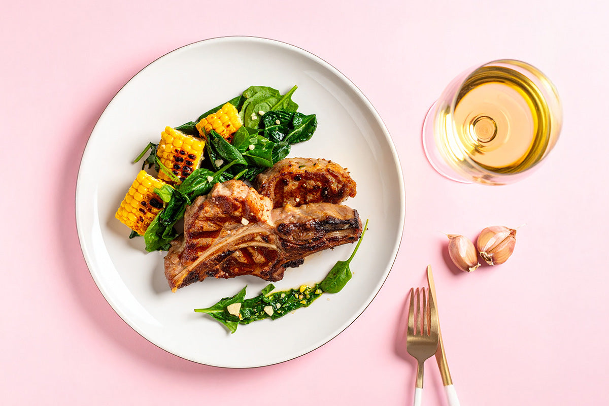 Gegrillte Lammkoteletts mit sautiertem Spinat und gegrillten Maiskolben auf weißem Teller, daneben Glas Weißwein und Knoblauchzehen