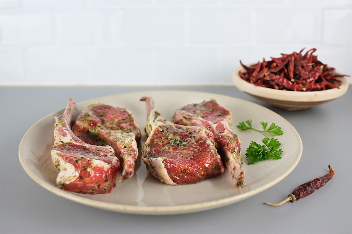 Vier marinierte Lammkoteletts auf einem weißen Teller mit Petersilienzweigen, daneben eine Schale mit getrockneten roten Chilischoten und eine einzelne Chilischote
