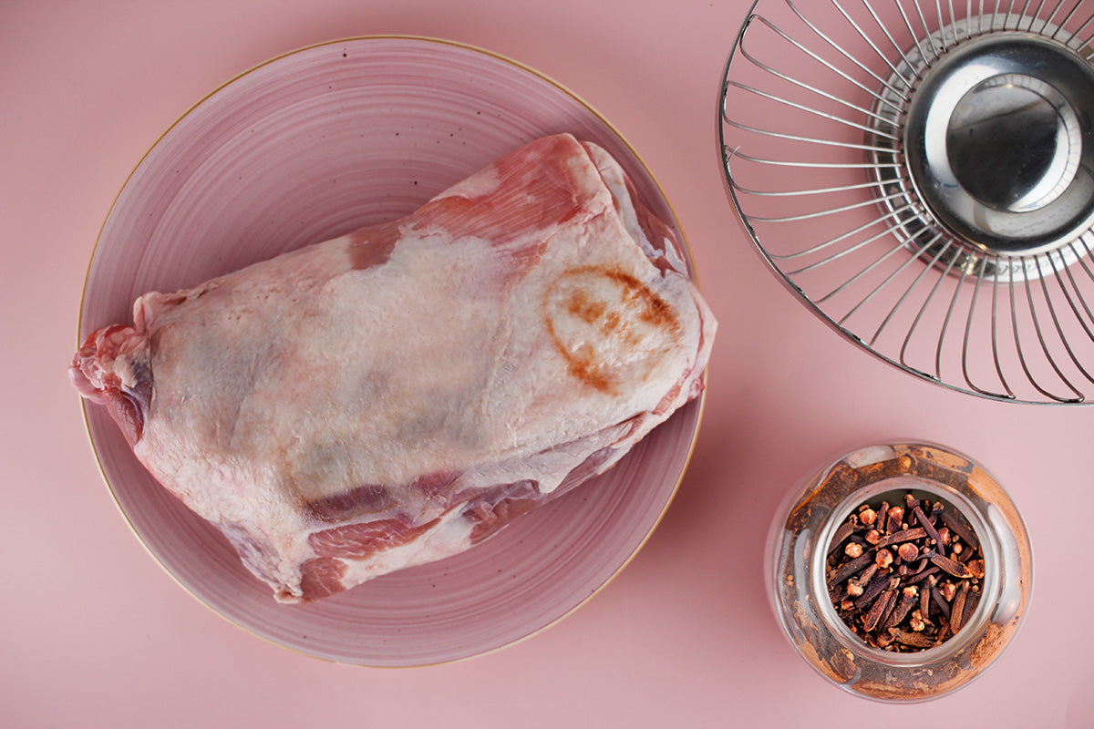 Rohe Lammschulter auf einem rosa Teller, daneben ein Glas mit Nelken und ein Metallkorb auf rosa Untergrund