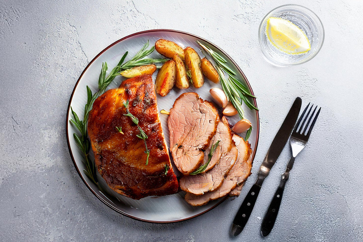 Gebratene Lammschulter mit Rosmarin, Knoblauch und Kartoffelspalten auf grauem Teller, daneben Messer, Gabel und Glas mit Zitronenscheibe