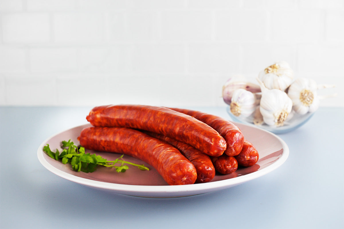 Rote Merguez-Würste aus Geflügelfleisch auf einem weißen Teller mit Petersilie, im Hintergrund Knoblauchknollen in einer Glasschale