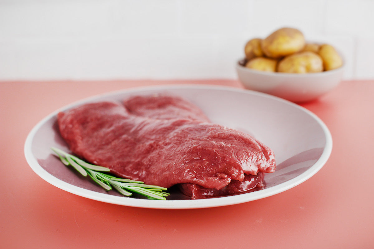 Zwei rohe Lammfilets auf weißem Teller mit Rosmarinzweig, im Hintergrund Schale mit kleinen Kartoffeln
