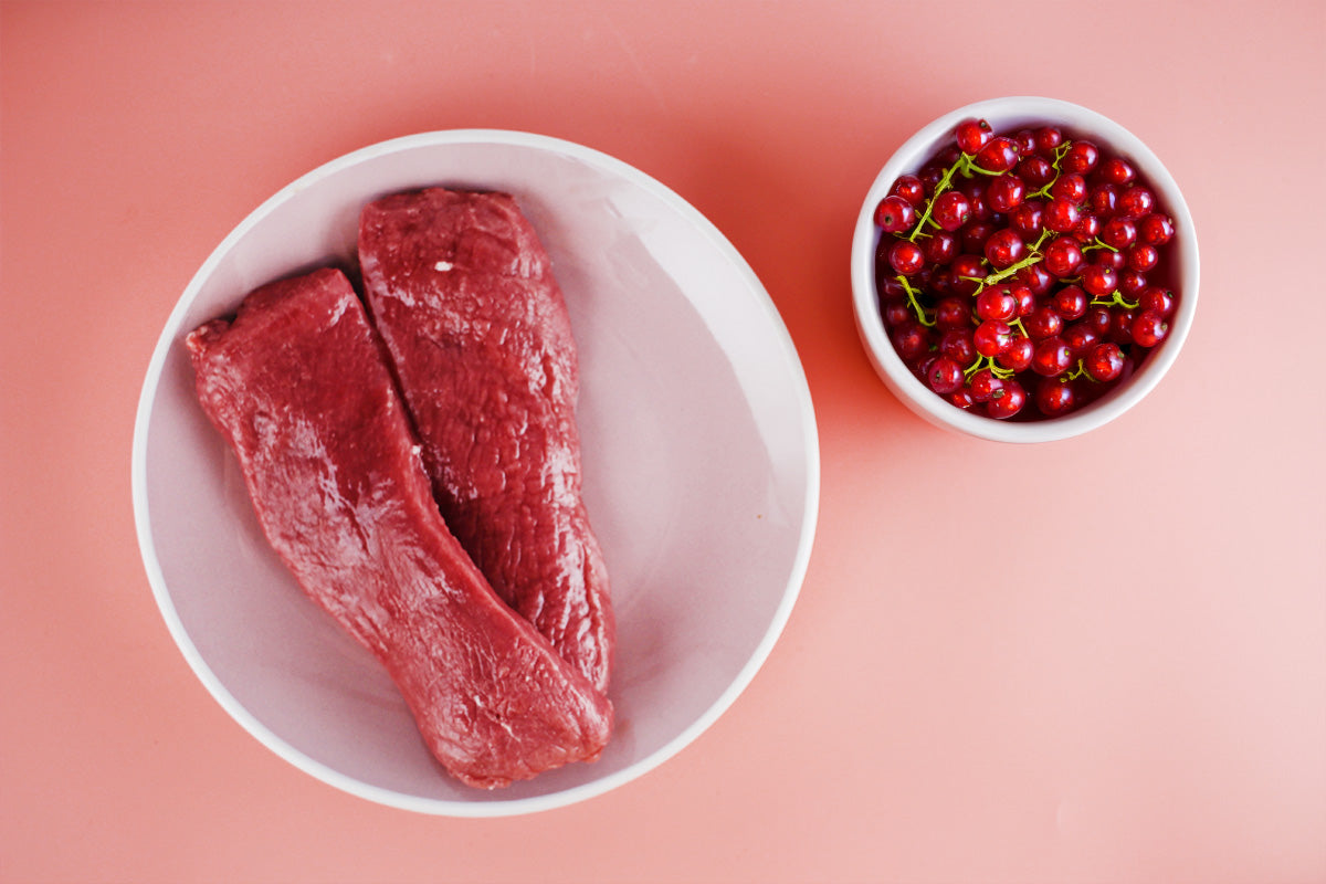 Zwei rohe Lammfilets in einer weißen Schüssel neben einer kleinen weißen Schüssel mit roten Johannisbeeren auf rosa Hintergrund