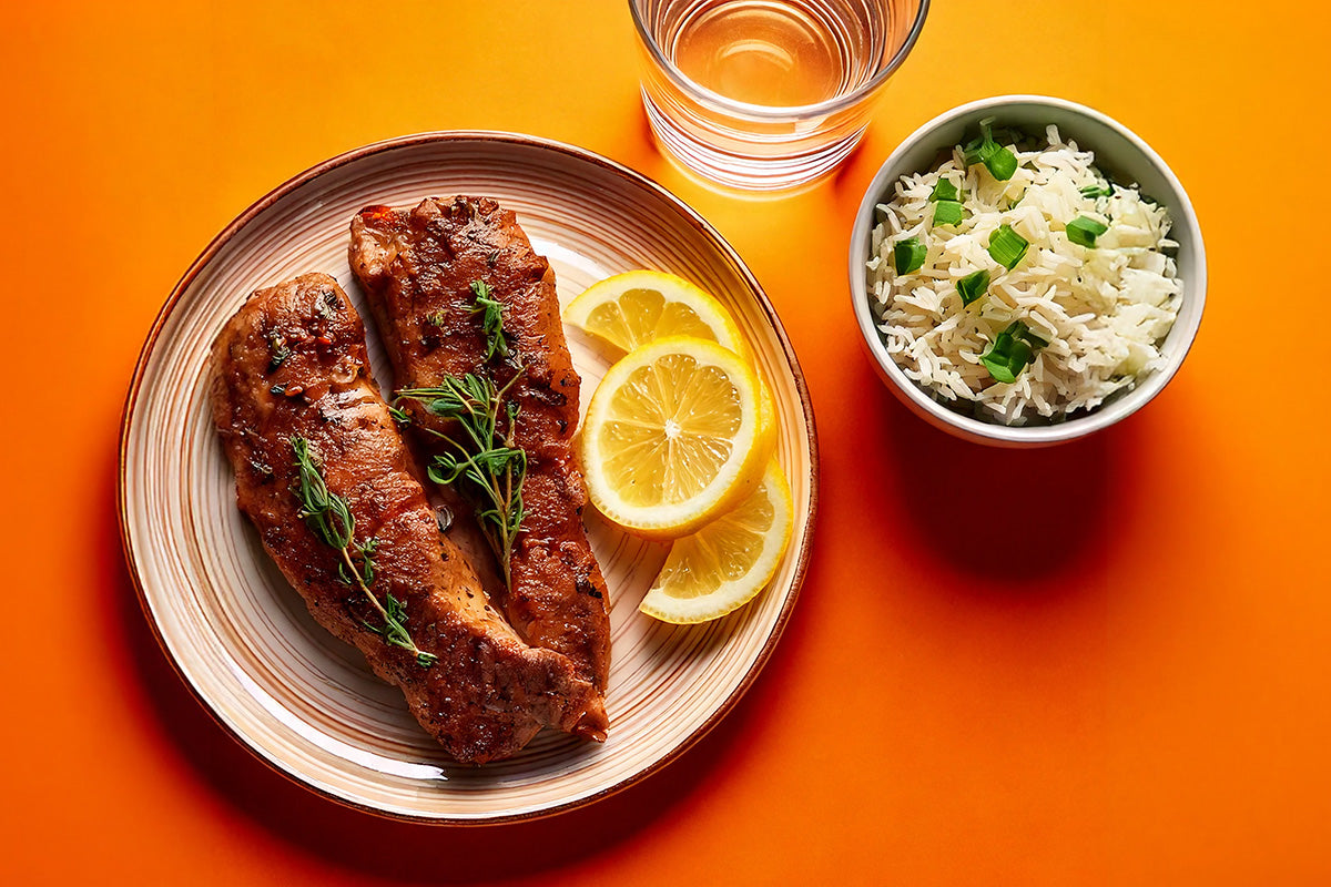 Zwei gebratene Lammfilets mit frischem Thymian und Zitronenscheiben auf einem Teller, daneben eine Schale Reis mit Frühlingszwiebeln und ein Glas Wasser auf orangefarbenem Hintergrund