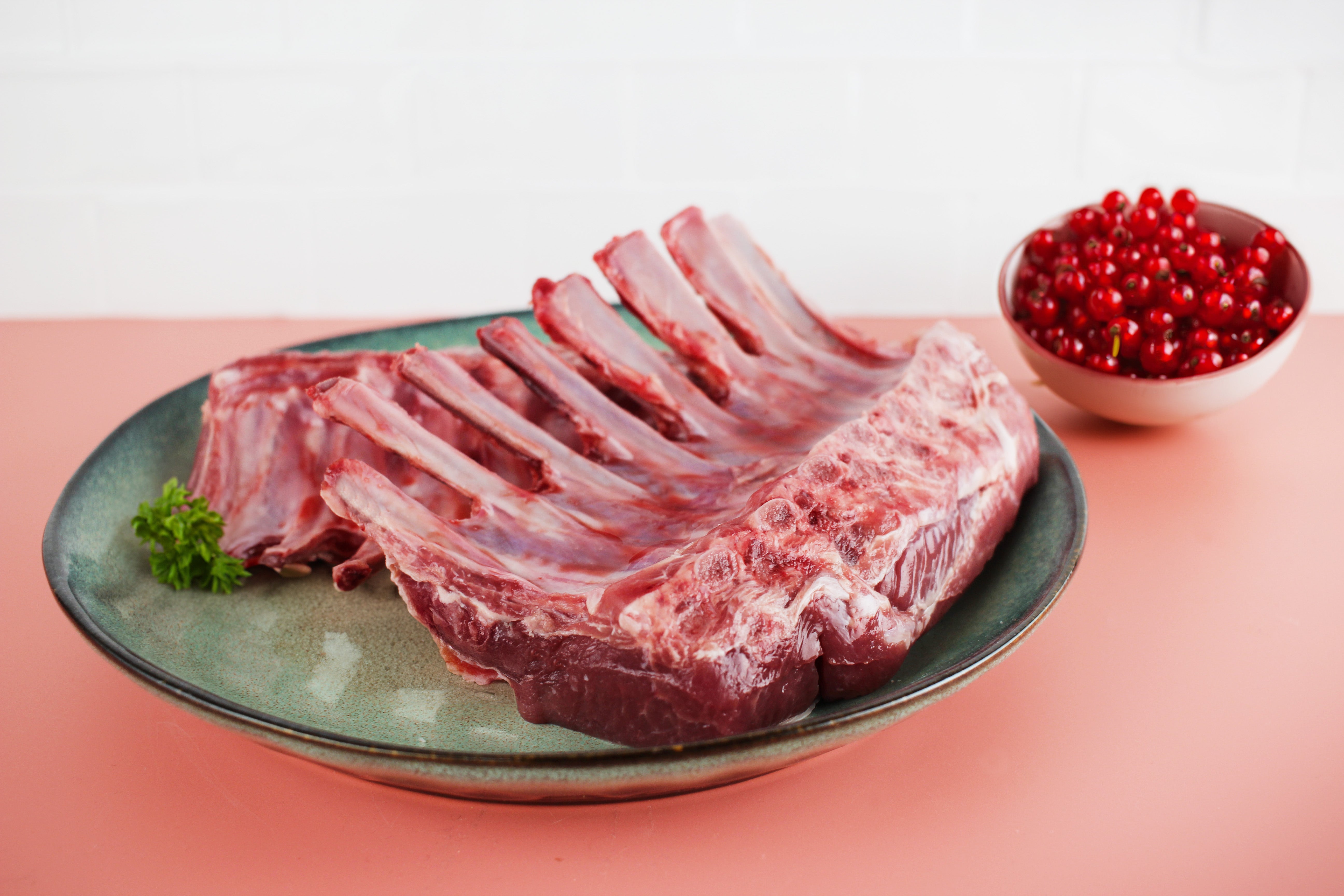 Zwei rohe Lammkarrees auf einem blauen Teller, garniert mit Petersilie, neben einer Schale mit roten Johannisbeeren auf rosa Hintergrund.