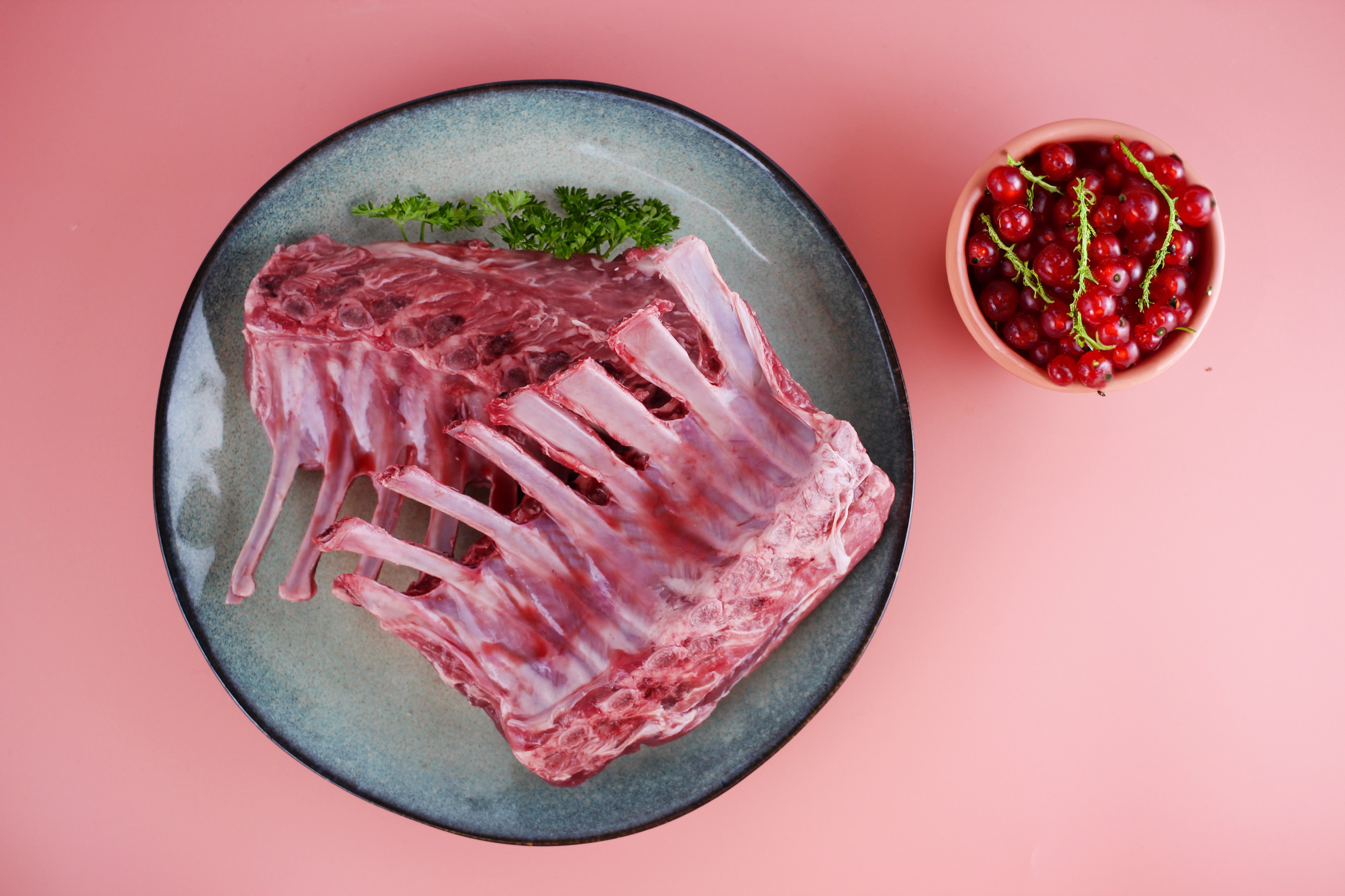 Zwei rohe Lammkarrees auf einem blauen Teller, garniert mit Petersilie, neben einer Schale mit roten Johannisbeeren auf rosa Hintergrund.