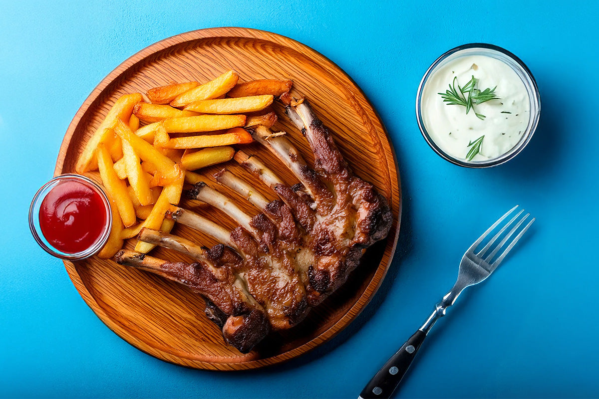 Auf einem Holzteller vor einem leuchtend blauen Hintergrund liegt gegrilltes Lammkarree, goldbraune Pommes, Ketchup und ein cremiger Dip und eine Gabel