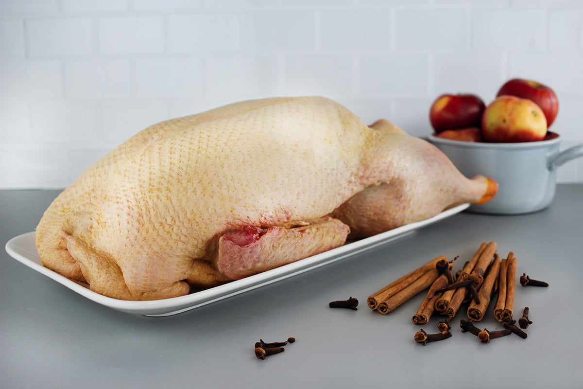 Rohe Gans auf weißem Teller, daneben Zimtstangen und Nelken, im Hintergrund Äpfel in einem grauen Topf