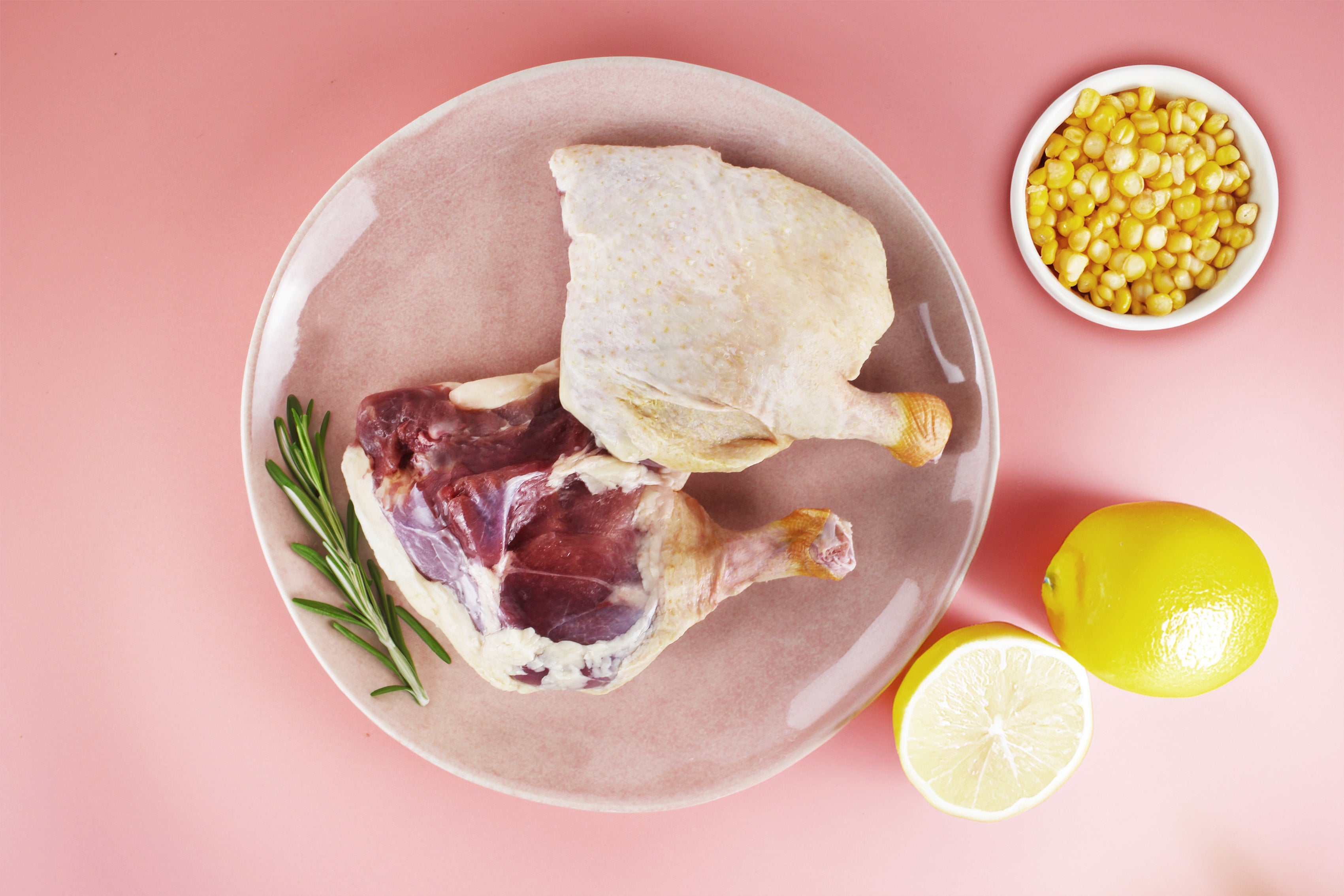 Zwei rohe Entenkeulen auf einem runden Teller, daneben ein Rosmarinzweig, eine halbe und eine ganze Zitrone sowie eine Schale mit Maiskörnern.