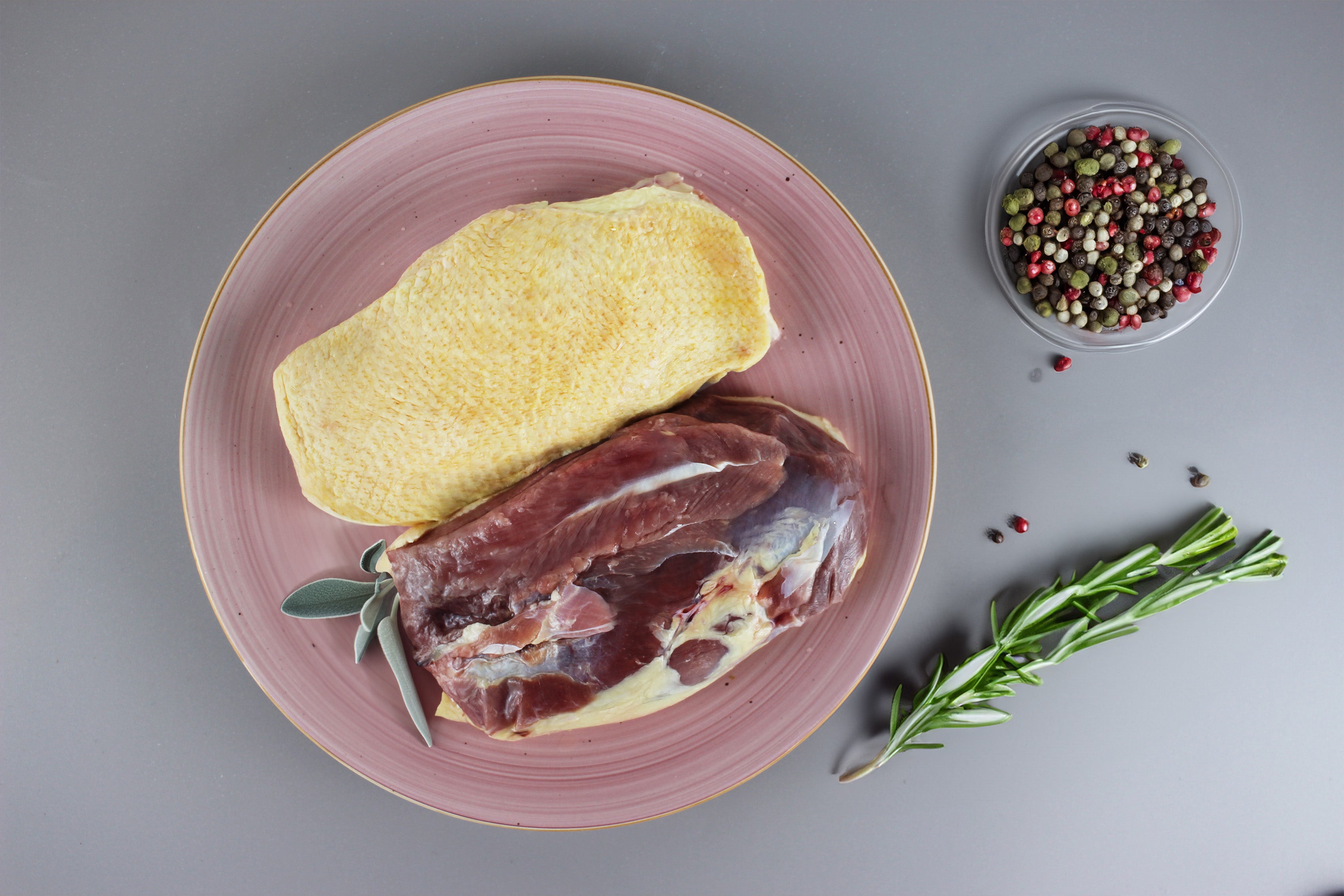 Rohe Gänsebrust mit Haut auf rosa Teller, daneben Rosmarinzweig und kleine Schale mit bunten Pfefferkörnern.