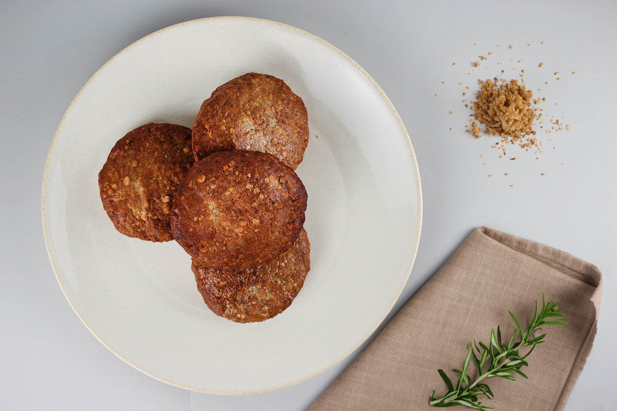 Drei gebratene Geflügelfrikadellen auf weißem Teller neben braunem Gewürz und Rosmarinzweig auf beigem Stoffserviette