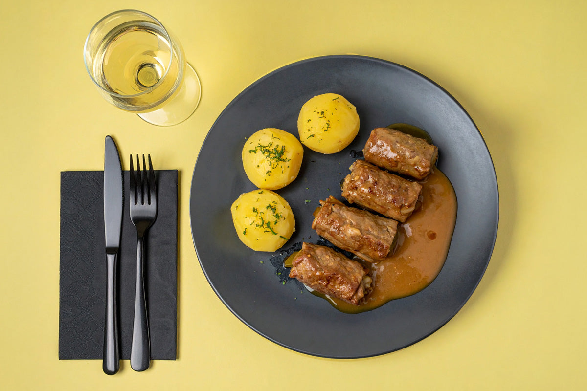 Dunkelgrauer Teller mit vier Putenrouladen in brauner Sauce und drei Salzkartoffeln, daneben Besteck auf schwarzer Serviette und ein Glas Weißwein