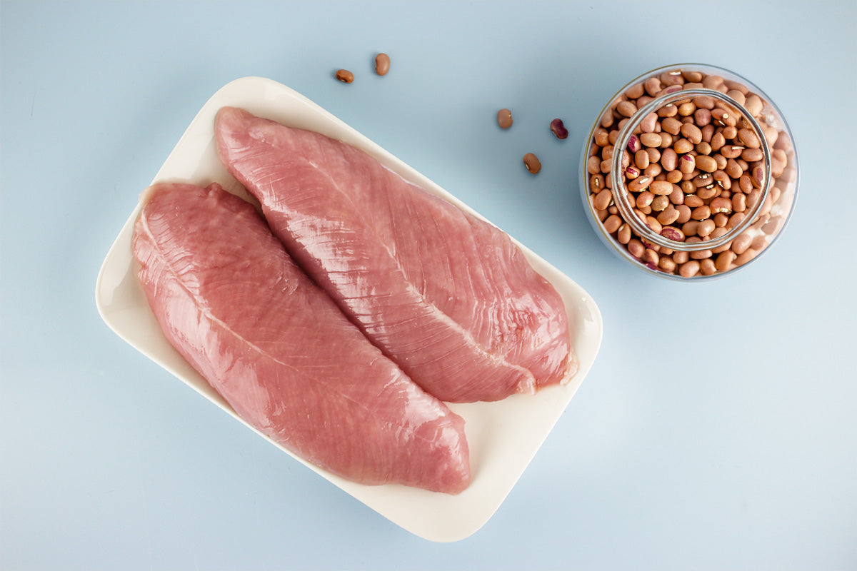 Zwei rohe Putenfilets auf weißem Teller neben Glas mit braunen Bohnen