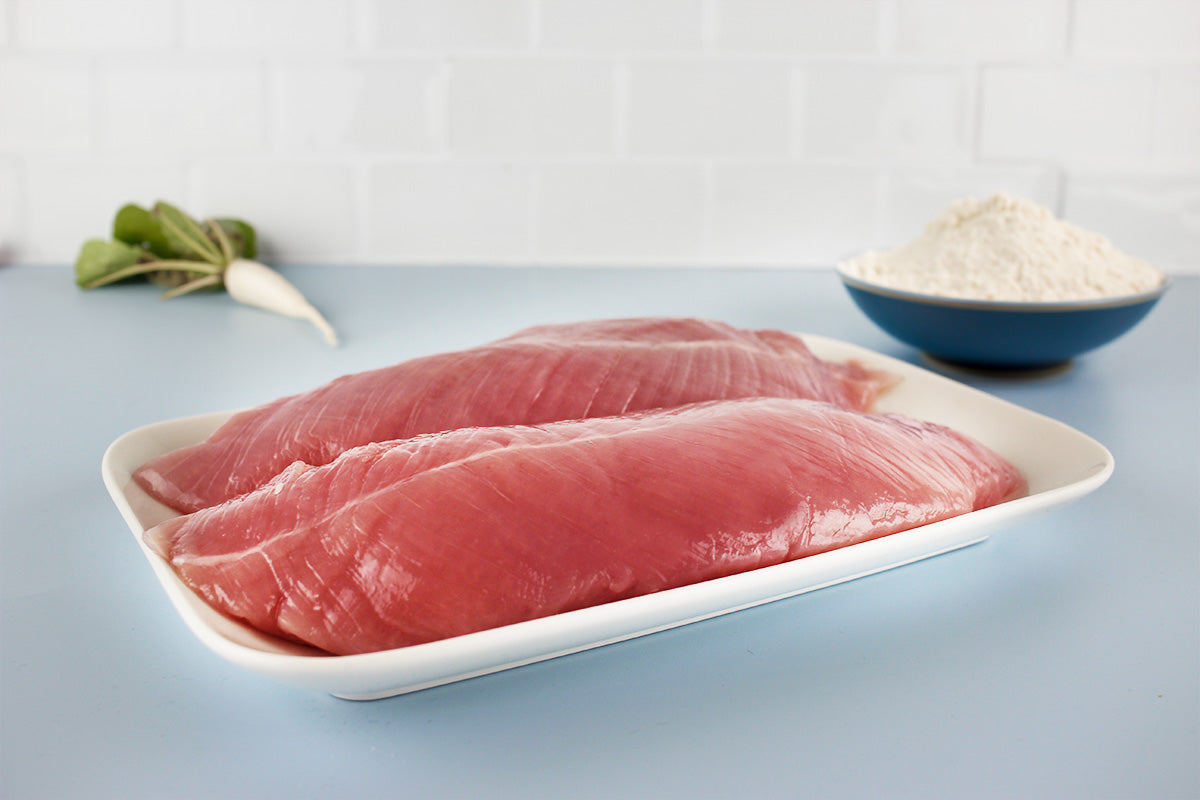 Zwei rohe Putenfilets auf weißem Teller, im Hintergrund eine Schüssel mit Mehl und ein Rettich